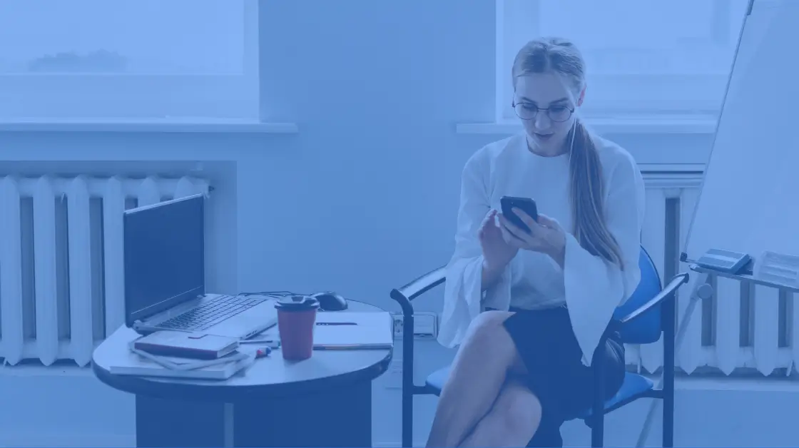 white woman in white top and black skirt sits looking at her phone, her laptop is on the table next to her